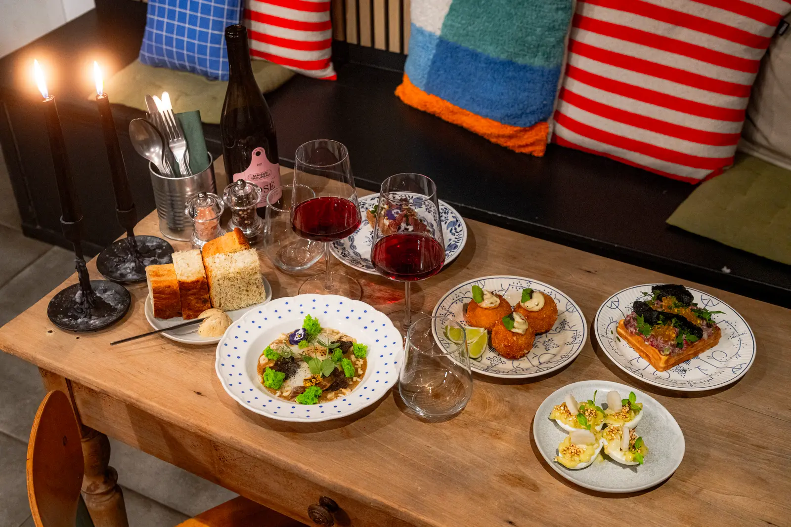 Une table en bois garnie de petites assiettes à partager, de pain, de deux verres de vin rouge et de bougies, devant une banquette aux coussins colorés.