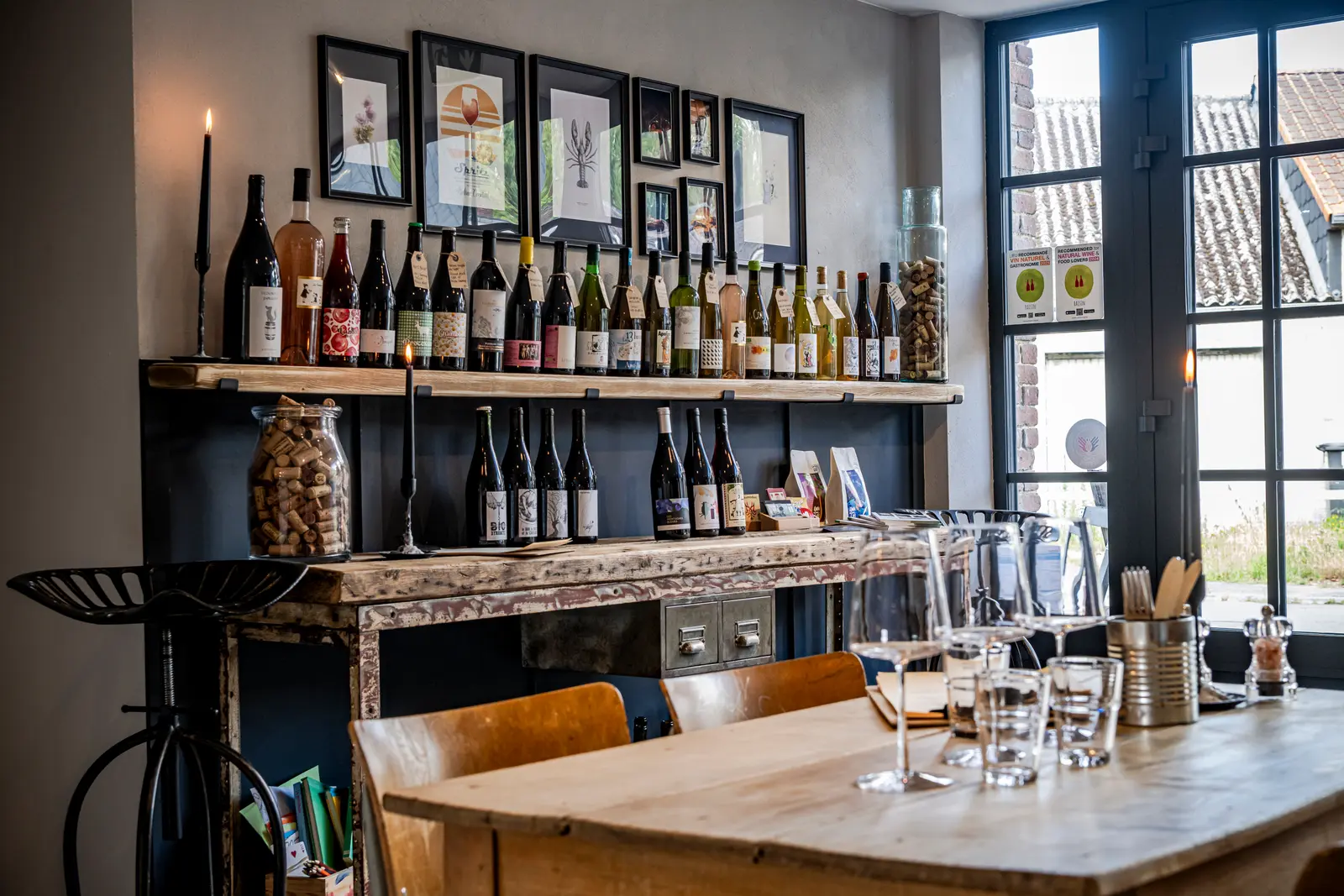 Une étagère en bois chargée de bouteilles de vin, sous des illustrations encadrées, à côté d'une grande fenêtre et d'une table dressée.